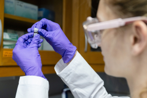 Out-of-focus researcher in protective laboratory gear and purple gloves holding a small vial in a chemistry lab.