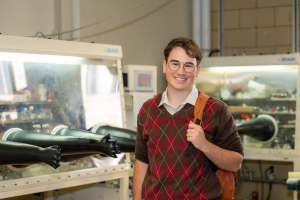 Nolan Cynkar stands in a lab with a backpack slung over one shoulder. He smiles at the camera.