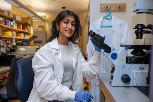 Female student wearing a lab coat and gloves sits at a workstation in a chemistry lab, preparing to use a microscope.