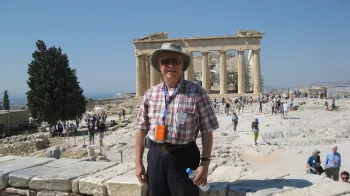 Victor Bearg stands in front of the Acropolis in Greece and smiles at the camera. He's wearing a plaid shirt, a hat and sunglasses.