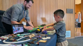 IDeATe Technical Specialist Cody Soska helps a young learner investigate the Plush Neuron. They both stand at a table with their hands on the Plush Neuron.