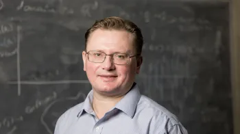 Sergey Nadtochiy stands in front of a blackboard with equations written on it. He smiles at the camera.
