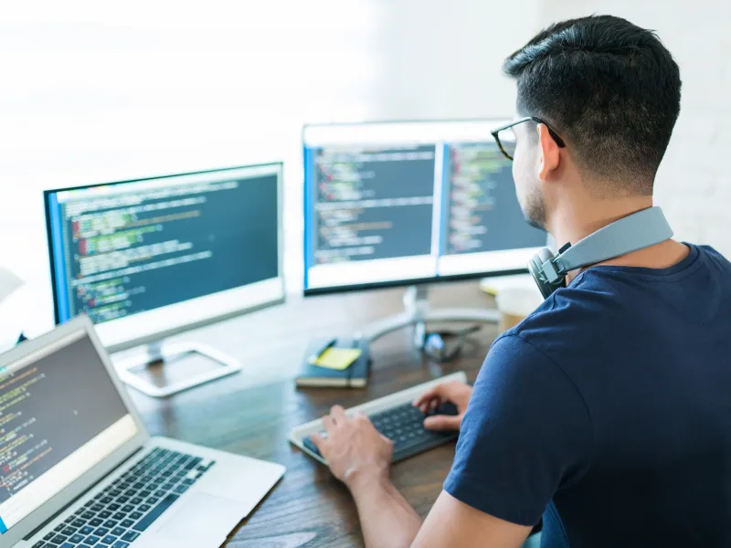 Back of young professional male at a desk with three computer monitors 