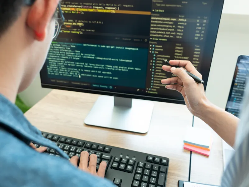 Image of code on a screen. One person's hands are on a keyboard and another person is pointing to the code on the screen. 