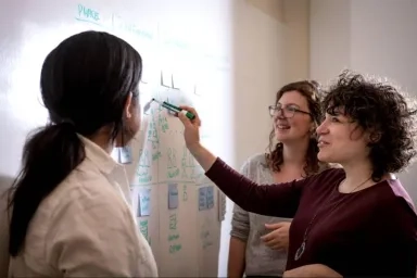 Two CMU continuing education students and a professor collaborate on a whiteboard covered with diagrams and sticky notes