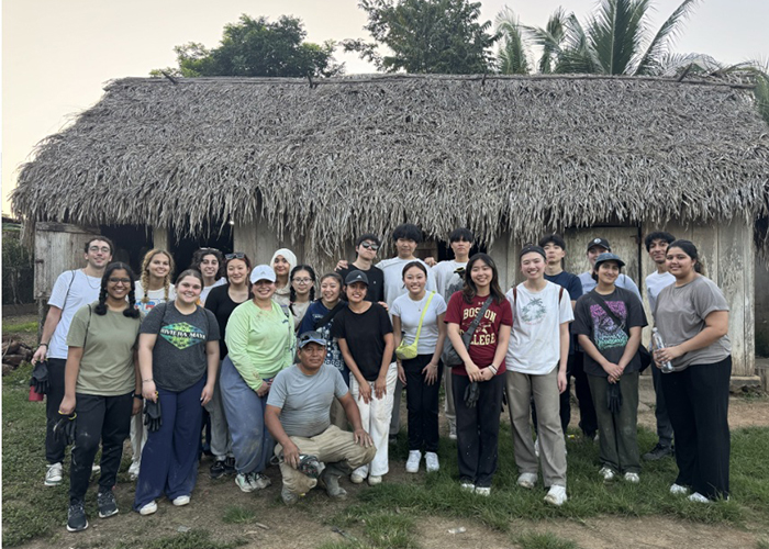a large group of CMU students in front of a structure with thatched roof