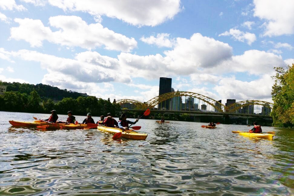 students kayaking on the river underneath Pittsburgh bridges and blue skies