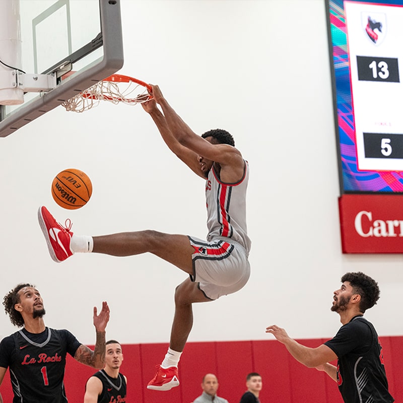 CMU Men's Basketball player dunks in mid-air during a competitive game in the Highmark Center for Health, Wellness, and Athletics