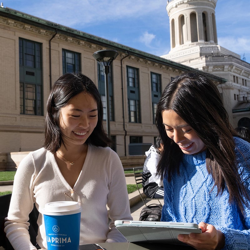 two students laughing at a table outside under a blue sky