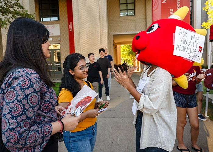 Activities Board (AB) member comically wears a dinosaur head costume with a sign taped to it reading, "Ask me about Activities Board," chats to interested students on the Cut at The FAIR 