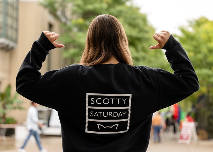 A student faces her back to the camera and points to the back of her shirt which reads, "Scotty Saturday"