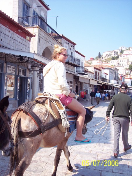 Kensee rides a camel through a narrow lane in Greece. The camel is guided by a man holding its harness.