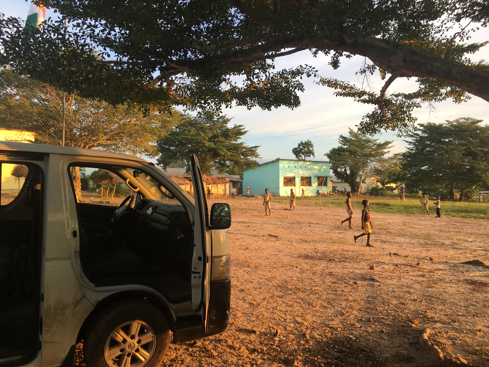 A van with the doors open is in front of a field and a small building. Kids are standing in the field.