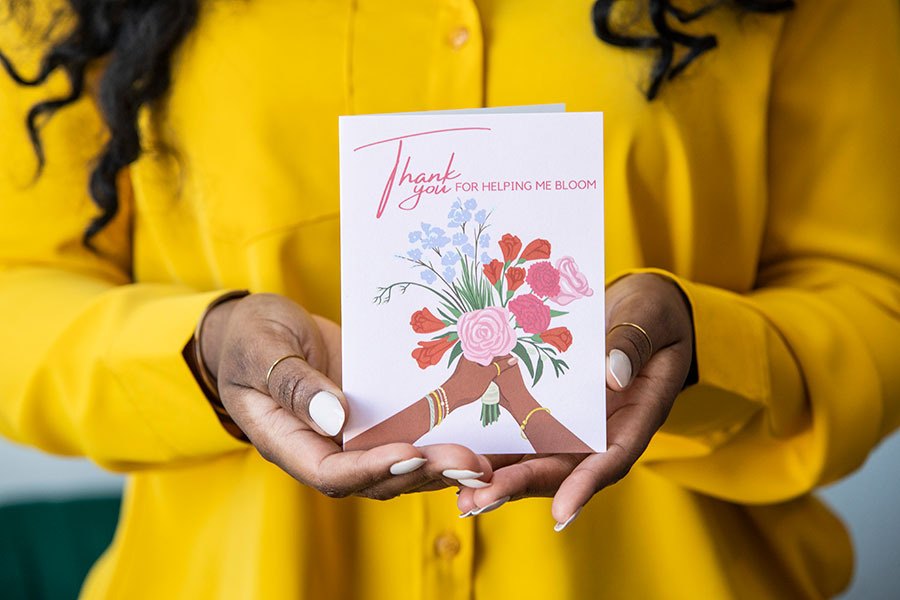 Asha Banks wearing a bright yellow suit holding one of her CheerNotes greeting cards toward the camera.