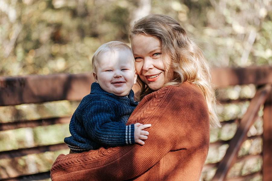 Angela wearing an orange sweater posing with her child in front of woods with autumn leaves.