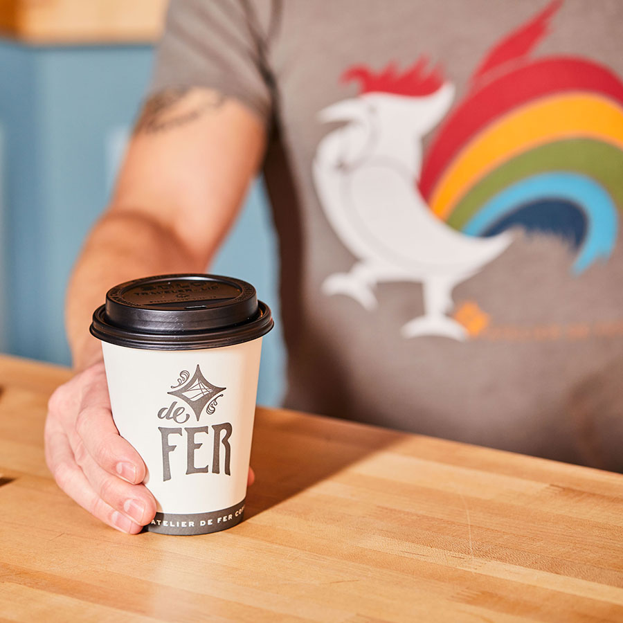 Man wearing rainbow rooster de Fer coffee logo t-shirt holding a cup of coffee over a wood counter