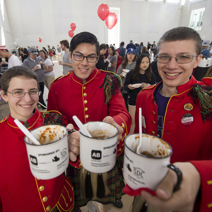 A group of smiling students dressed in band uniform eating chili from mugs.