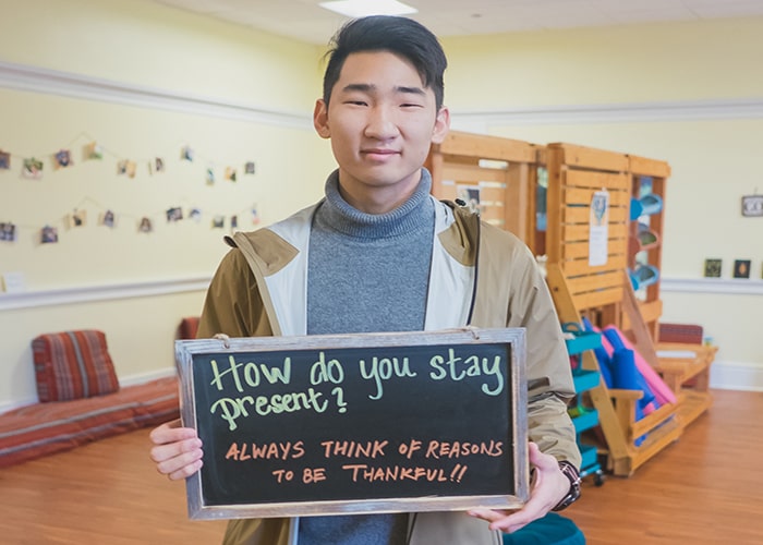 Student holds up chalkboard sign that reads "How do you stay present? Always think of reasons to be thankful!" in the Mindfulness Room