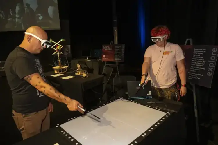 Two people wearing smart glasses, interacting with a table using controllers.